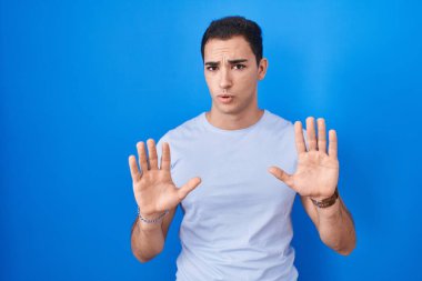 Young hispanic man standing over blue background moving away hands palms showing refusal and denial with afraid and disgusting expression. stop and forbidden. 