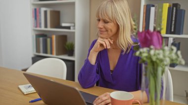 Gülümseyen sarışın bir kadın evinde keyfine bakıyor. Dizüstü bilgisayarı, çiçekleri ve masanın üstünde bir not defteri var..