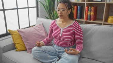 Sükûnetli İspanyol bir kadın, gri bir kanepede, renkli yastıklarla çevrili, sükunet ve sükunet içinde meditasyon yapıyor..