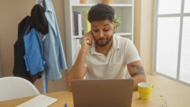 Düşünceli, Afro-Amerikan bir adam evde çalışırken bilgisayarını kullanıyor ve telefonla konuşuyor..