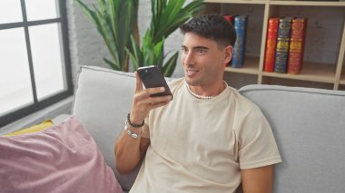 Gülümseyen İspanyol bir adam, rahat bir oturma odasında, elinde akıllı bir telefonla, bir kanepede öylesine oturuyor..