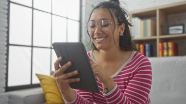 Gülümseyen genç İspanyol kadın kitaplığın arka planında, modern oturma odasında tablet kullanıyor..