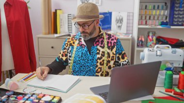 Şık bir adam renkli tekstil, dikiş makinesi ve moda çizimleriyle çevrili canlı bir terzi dükkanında eskizler çiziyor..