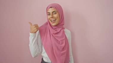 Tesettürlü gülümseyen genç İspanyol kadın pembe izole edilmiş bir arka plana başparmağıyla işaret ediyor..