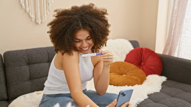 Gülümseyen genç bir kadın hamilelik testi ile evde selfie çeker, neşe ve beklenti ifade eder..