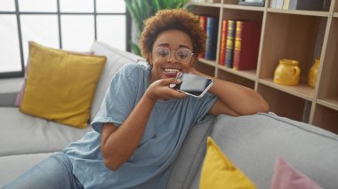 Gülümseyen siyah kadın evde akıllı telefonuyla kanepede dinleniyor.