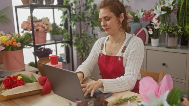 Kırmızı önlüklü genç bir kadın çiçek buketleriyle dolu bir çiçekçinin içinde dizüstü bilgisayarında güller düzenliyor..