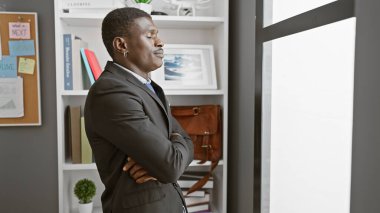 Confident african american man with arms crossed standing in a modern office setting. clipart