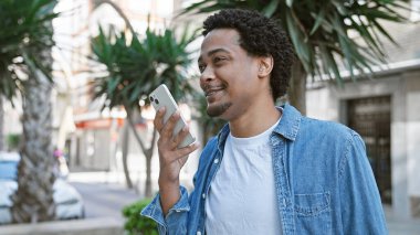 Gülümseyen, kıvırcık saçlı, caddede akıllı telefon kullanan Afro-Amerikan bir adam.