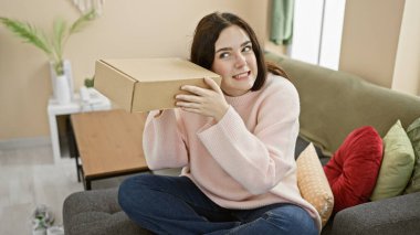 Curious young woman listens to a mystery cardboard box while sitting comfortably in her modern living room. clipart