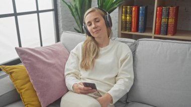 Rahat genç bir kadın kulaklıkla müzik dinlemekten zevk alır. Elinde akıllı bir telefon, rahat bir oturma odasındaki kanepede uzanır..