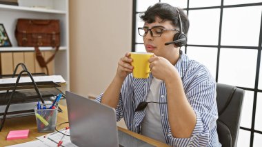 Kulaklık takan genç bir adam modern bir ofiste masasında kahve içiyor, üretkenliği ve gündelik profesyonelliği somutlaştırıyor..