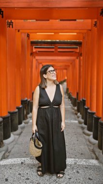 Gözlüklü kendine güvenen İspanyol güzeli, kulaktan kulağa sırıtan, Kyoto 'daki Fushimi tapınağındaki turuncu kırmızı torii tüneline huşu içinde bakan.!