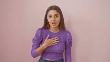 Elleri göğsünde, pembe bir arka planda mor bir süveter içinde duran, şaşırmış, genç İspanyol bir kadın..