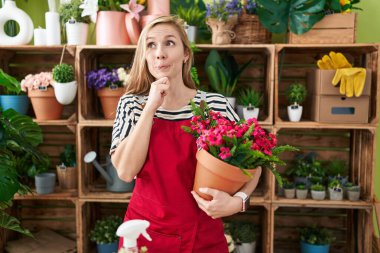Çiçekçi dükkanında çalışan beyaz bir kadın elinde bitkiyle ciddi bir yüz ifadesi olan eli çenesinde soru soran, kafa karıştırıcı bir fikir hakkında düşünceli bir kadın. 