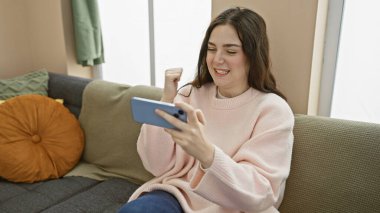 Pembe kazaklı beyaz bir kadın, bir apartman dairesinde akıllı telefonuyla kazandığı zaferi coşkuyla kutluyor..