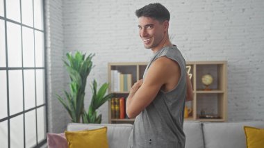 Kolsuz tişörtlü gülümseyen genç bir İspanyol. Gri bir kanepesi ve beyaz tuğlalı duvarı olan modern bir oturma odasında duruyor..
