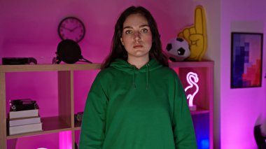 A young woman wearing a green hoodie stands in a colorful gaming room with neon lights at night. clipart