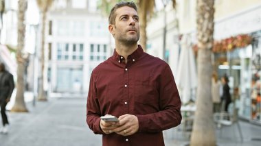 Yakışıklı İspanyol sakallı adam güneşli şehir caddesinde akıllı telefon kullanıyor..