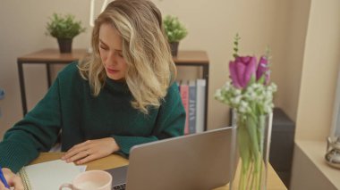 Evde dizüstü bilgisayarıyla çalışan odaklanmış sarışın bir kadın, iç dekorasyonla çevrili, serbest yaşam tarzını somutlaştırıyor..