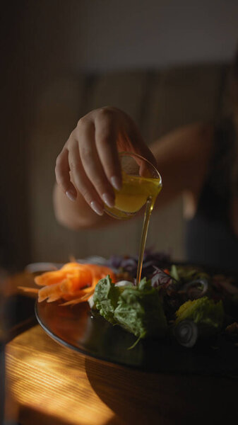 A young woman pours dressing on a salad in a modern cafe setting, suggesting a healthy dining lifestyle.