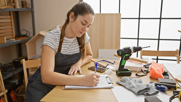 Сосредоточенная женщина пишет заметки в деревообрабатывающей мастерской, окруженной инструментами и техникой безопасности.