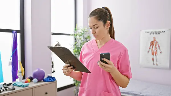 一名穿着粉红刷子的专注的年轻女子 带着治疗设备 检查了康复中心内部的剪贴板和智能手机 图库照片