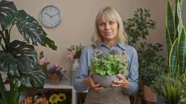 Sarışın bir kadın, arka planda çeşitli yeşillikler olan yemyeşil bir çiçekçide saksı tutuyor..