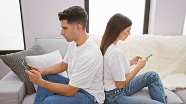 Bir çift arka arkaya oturma odalarında oturuyor, akıllı telefonlarıyla meşgul, modern kapalı bir yaşam tarzını yansıtıyor..