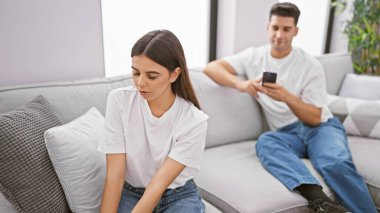 Bir erkek telefonunu kullanırken bir kadın üzgün görünür. Modern bir oturma odasında ilişki sorunlarına işaret eder..
