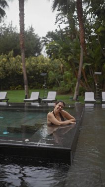 Gülümseyen genç bir kadın, bereketli tropikal yeşilliklerle çevrili bir bale merkezindeki açık havuzda huzurlu bir anın tadını çıkarıyor..