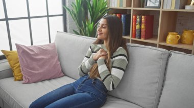 Elleri göğsünde genç bir İspanyol kadın, rahat bir oturma odasında, bir kanepede huzur içinde oturuyor, sakin ve rahat bir şekilde yayılıyor..