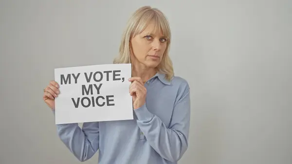 stock image A middle-aged woman with blonde hair, standing against a white backdrop, holds a sign about voting rights.