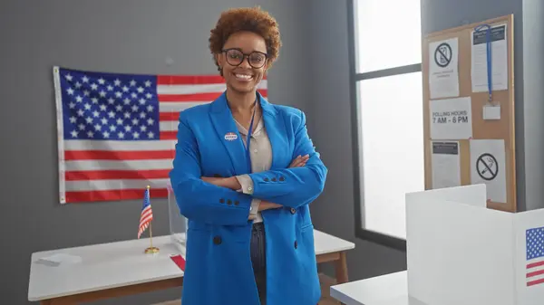 Stock image Confident african american woman smiling in a voting center with an american flag, portraying democracy and civic duty.