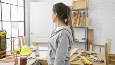 Etrafı ahşap işçiliği aletleri ve ekipmanlarla çevrili bir marangoz atölyesinde duran kendine güvenen genç bir İspanyol kadın..