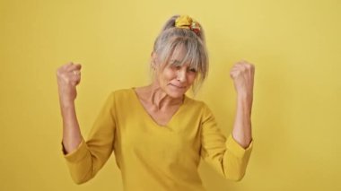 Gri saçlı at kuyruklu neşeli orta yaşlı kadın zaferi kutluyor! Heyecanlı kazanan başarı çığlıkları atıyor, sarı arka planda izole edilmiş..