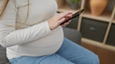 Hamile İspanyol kadın evde otururken akıllı telefon kullanıyor, modern annelik yaşam tarzını örnek alıyor..