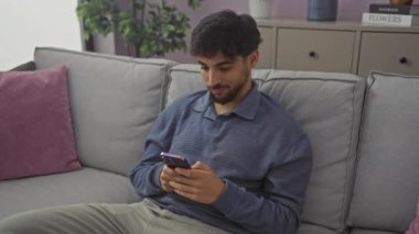 Şok olmuş genç Arap adam, elinde eliyle ağzını kapatıyor. UFO gözlü bir sürpriz, evdeki kanepede oturuyor. Akıllı telefondan şok edici haberler izledikten sonra korkmuş ve nutku tutulmuş.