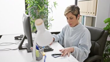 Kısa sarı saçlı, gülümseyen bir kadın modern ofis ortamında akıllı telefonunu kullanarak gündelik bir andan zevk alır..