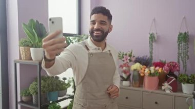 Gülümseyen sakallı bir adam çiçekçi dükkanında selfie çekiyor. Etrafı renkli çiçekler ve ev bitkileriyle çevrili..