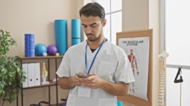 Tıbbi üniformalı genç sakallı bir adam arka planda rehabilitasyon ekipmanları olan bir klinikte akıllı telefon kullanıyor..