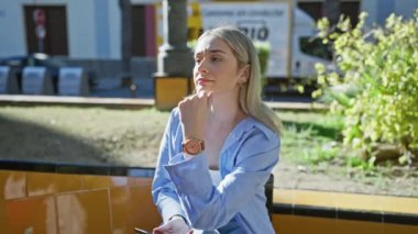 Sarışın genç bir kadın, elinde akıllı bir telefonla, güneşli bir parkta otururken kol saatini kontrol ediyor..