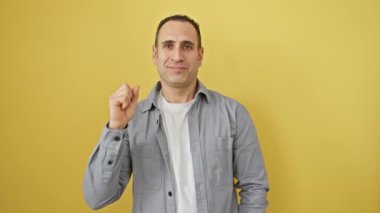Neşeli, genç İspanyol bir adam iki parmağıyla işaret ediyor, sarı izole bir arka planda duruyor, tişört giyiyor ve mutlu bir şekilde gülümsüyor.
