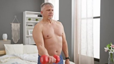 Olgun, üstsüz bir adam evde dambıllarla egzersiz yapıyor, spor yapıyor ve sağlığını gösteriyor..