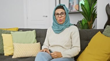 Orta yaşlı bir kadın tesettür takıyor modern bir oturma odasında rahat bir şekilde oturuyor gri bir kanepede renkli yastıklarla.