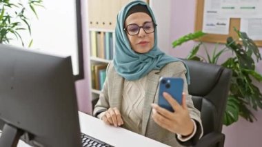 Başörtüsü takan gülümseyen bir kadın modern ofiste akıllı telefonuyla selfie çekiyor..