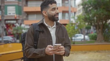 Sakallı, gülümseyen genç adam şehir parkında akıllı telefon kullanıyor..