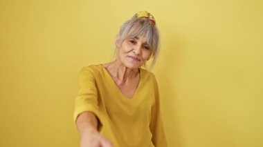 Sıcak, pozitif ve orta yaşlı, gri at kuyruklu İspanyol kadın izole edilmiş sarı arka planda gülümseyen, misafirperver bir el sıkışma sunuyor. İş hayatında başarı!