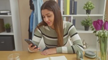 Genç İspanyol kadın evinde, rahat bir oturma odasında çiçeklerle ve modern dekorla akıllı telefonuyla geziniyor..