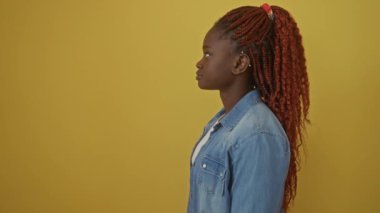 Kendine güvenen Afro-Amerikalı kadın doğal bir şekilde duruyor, kot gömlek giyiyor, dost canlısı bir gülümseme ile yan yana bakıyor, sarı arka planda izole edilmiş.